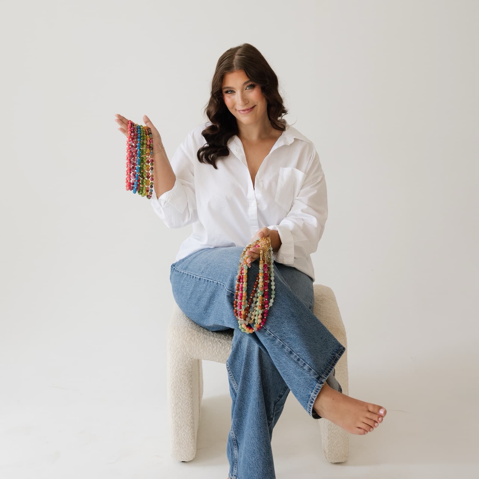 Taylor smiling on a boucle stool in a white shirt and jeans, holding strands of colorful beaded necklaces